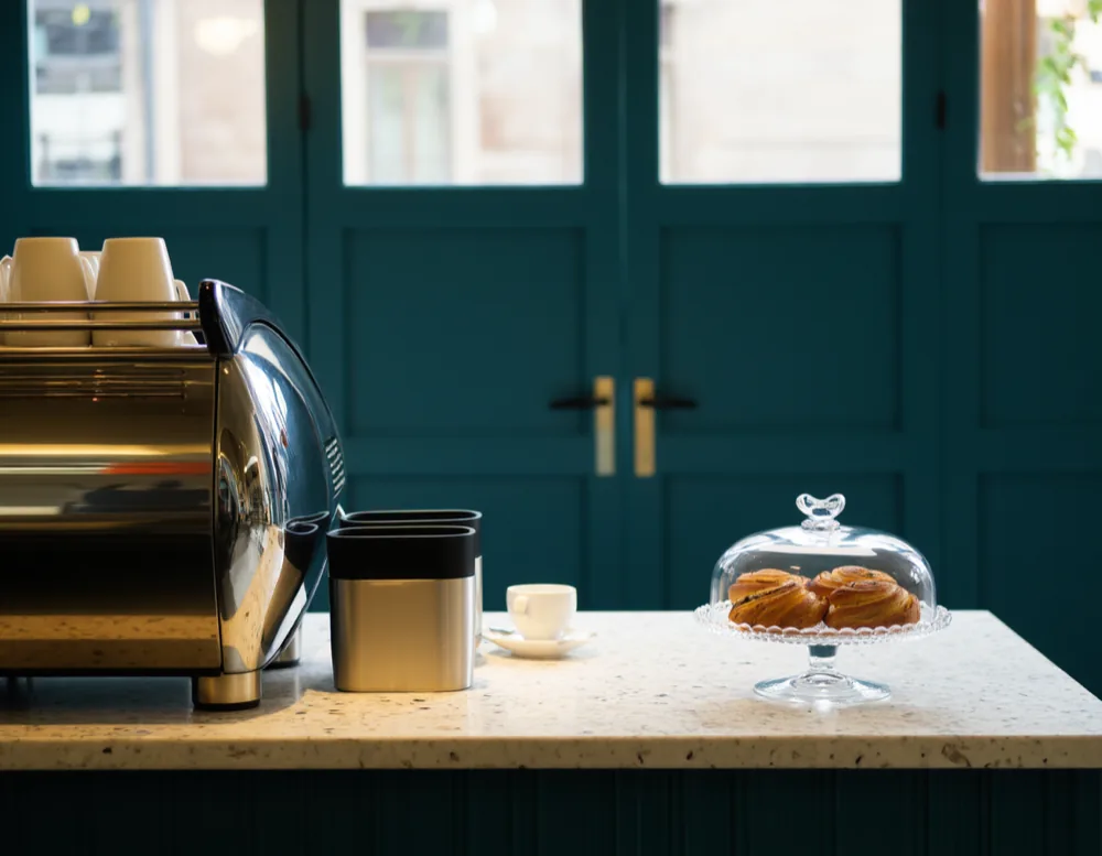 The counter: a chrome espresso machine, stacked cups and cinnamon buns under a glass dome on a terrazzo top, against deep teal panelling.