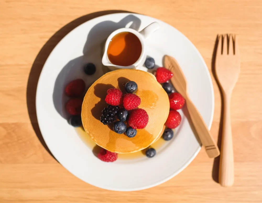A kids' pancake seen from above: one pancake with berries, a small jug of maple syrup and a wooden fork on an oak table.