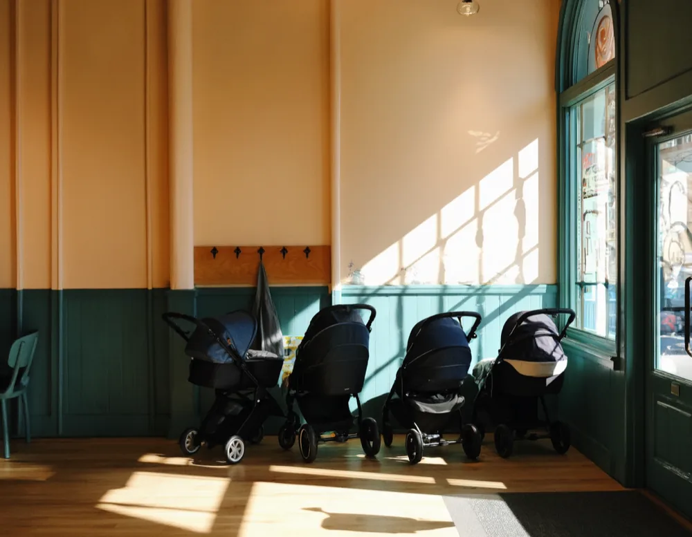 Four prams parked side by side under a tall sunlit window in the front hall, with teal panelling behind them.