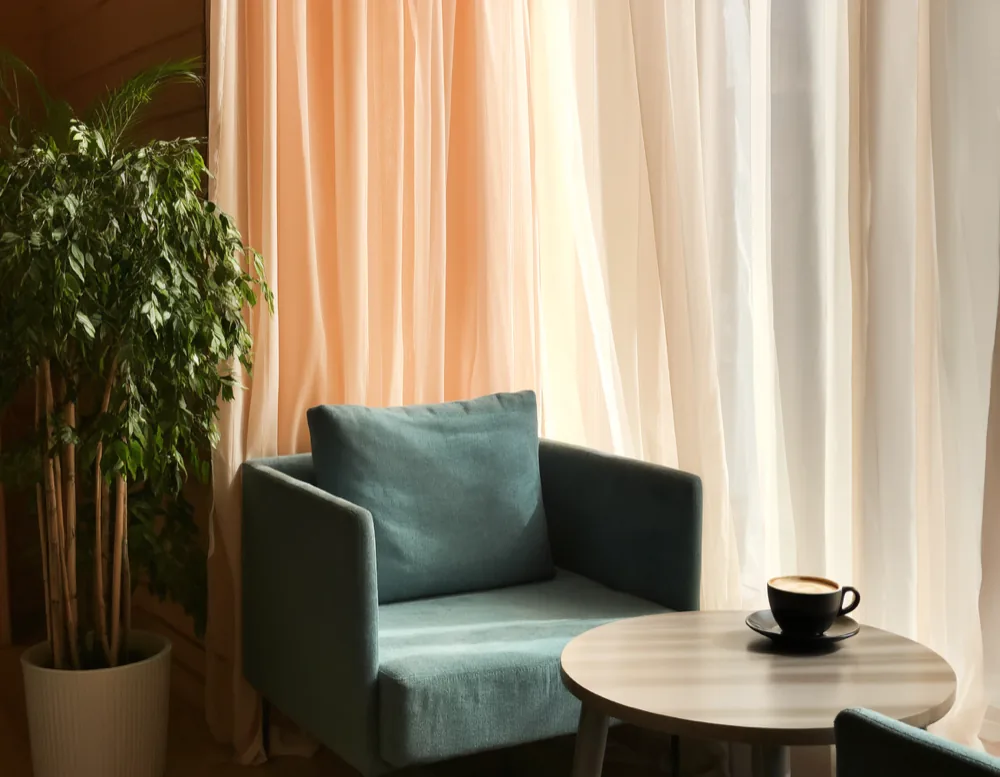 A quiet corner of the café during a Ruhezeit block: a teal armchair, a small round table with a single coffee, and daylight diffused through a linen curtain.